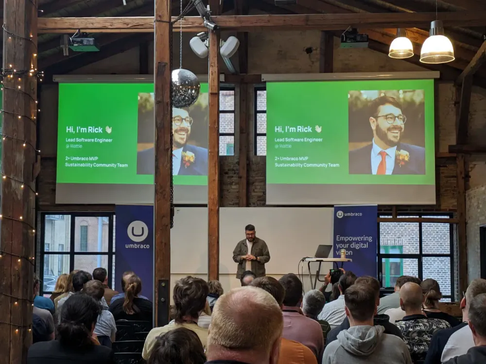 A full conference room with two large projector screens on the back wall, featuring a bright green slide, Rick's picture and text introducing Rick. He is stood on the stage at the bottom.
